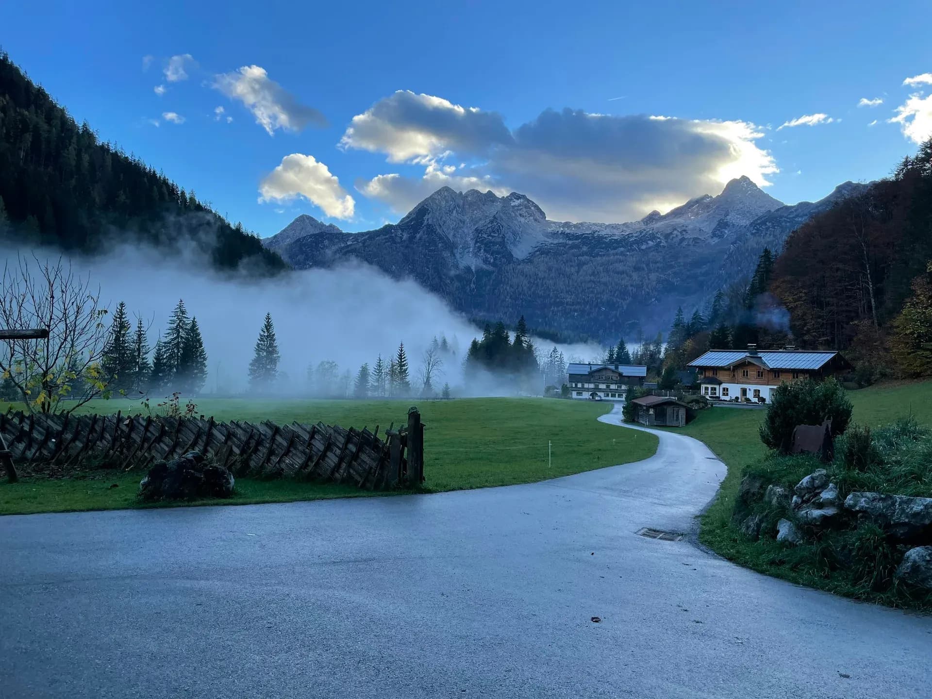 Herbstnebel über einer Wiese im Tal, dahinter Berge und ein Bauernhaus