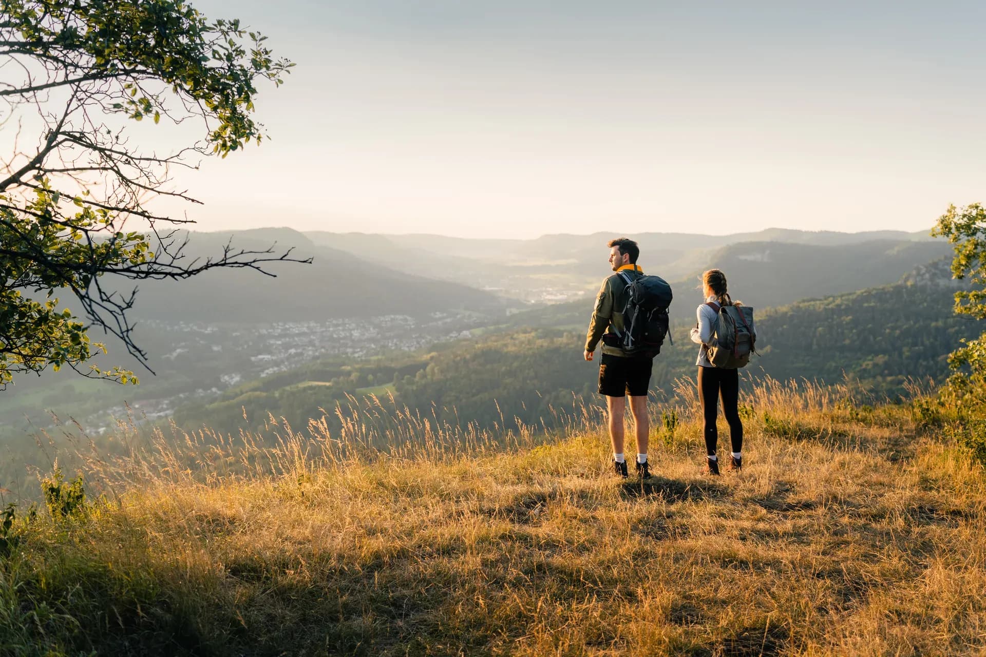 Zwei Wandernde mit Rucksack stehen im Morgenlicht auf einer herbstlichen Wiese über einem Tal